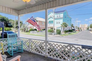 A covered porch with a view of a residential street, a rocking chair, and an American flag. - Ocean Gem (North Myrtle Beach)