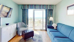 Ein Zimmer mit einer Couch, Sesseln, Hocker, Fernseher und Blick ins Freie durch eine Schiebetür.