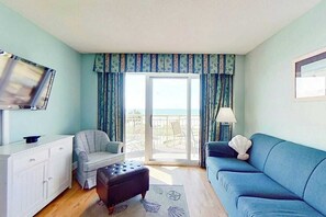 Ein Zimmer mit einer Couch, Sesseln, Hocker, Fernseher und Blick ins Freie durch eine Schiebetür.