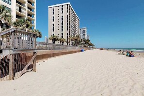 A beach with a wooden walkway, palm trees, and a high-rise building in the background. - Oceanfront Arcadian II  8D (Myrtle Beach)