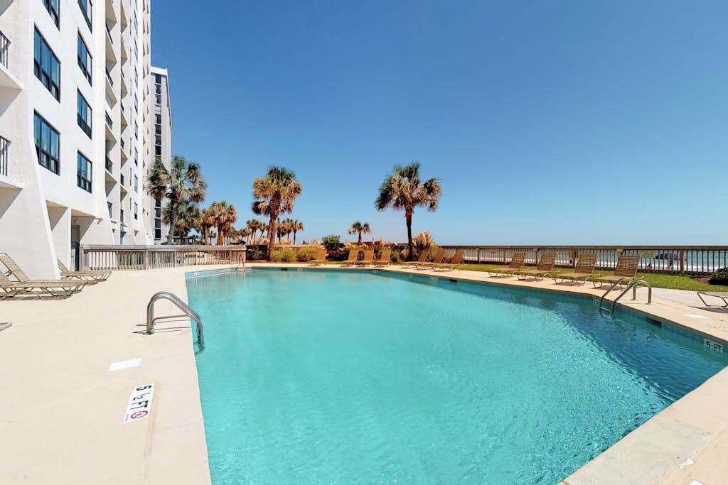 A swimming pool with lounge chairs and palm trees, adjacent to a white building.
