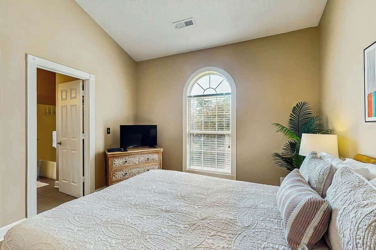 Un dormitorio con una cama grande, un televisor sobre una cómoda, una ventana con persianas y una puerta que da a otra habitación.