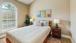 Una camera da letto con un letto grande, una finestra con persiane, un comodino con una lampada e una parete con tre opere d'arte incorniciate.