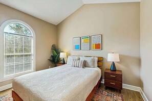 Ein Schlafzimmer mit einem großen Bett, einem Fenster mit Jalousien, einem Nachttisch mit Lampe und einer Wand mit drei gerahmten Kunstwerken.