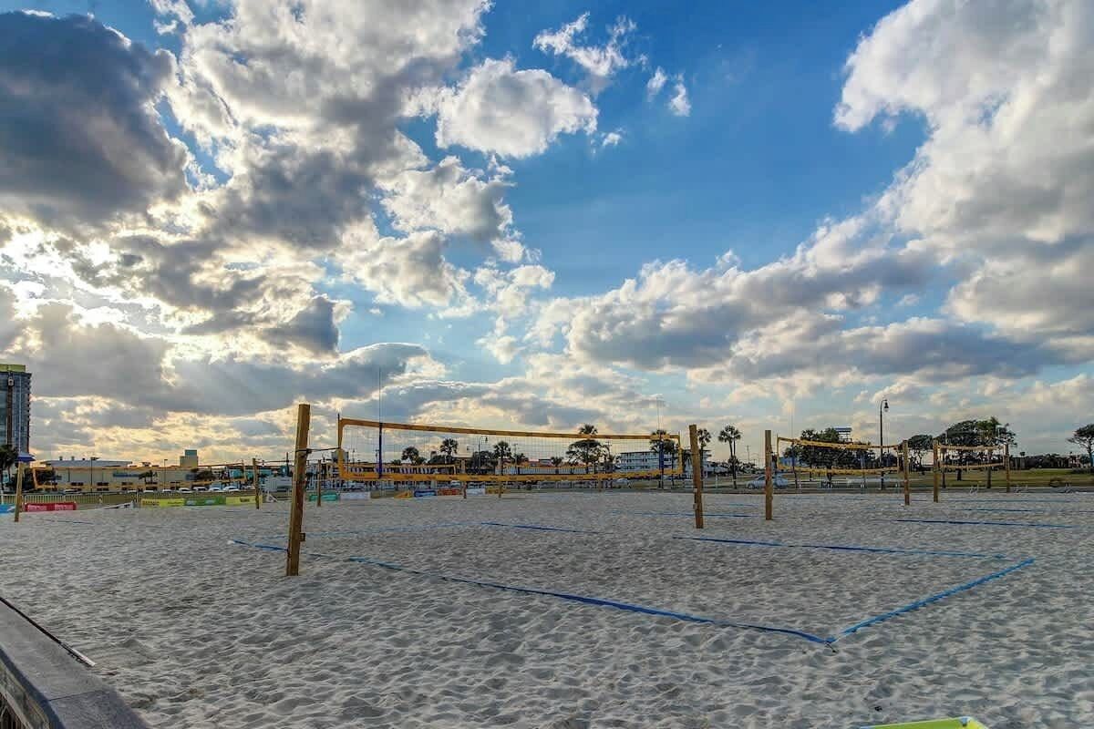 A beach volleyball court with net and boundary lines, surrounded by sand and palm trees under a partly cloudy sky.