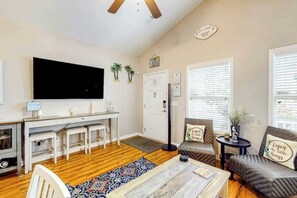 A living room with a TV, a dining table, and two chairs. There is a desk with a computer and a chair. The room has a wooden floor and a window with blinds. - Palm Tree Paradise Beach Cottage (Myrtle Beach)