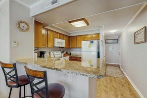 A kitchen with wooden cabinets, a microwave, a refrigerator, and a bar area with stools. - Pelicans Watch 106 On the Beach (Myrtle Beach)