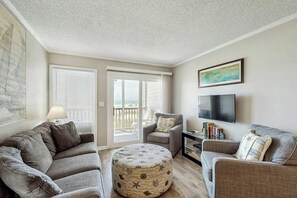 A living room with a sofa, ottoman, and armchairs, a TV, and a balcony with a view. - Pelicans Watch 106 On the Beach (Myrtle Beach)
