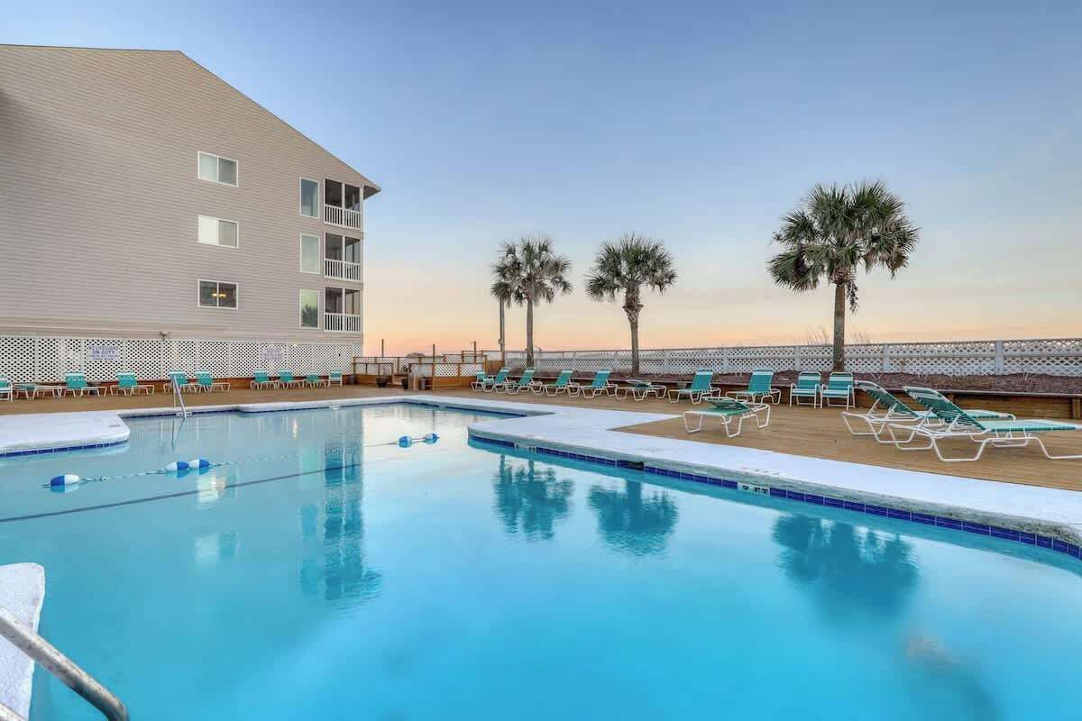 A swimming pool with lounge chairs and palm trees, surrounded by a building with multiple balconies.