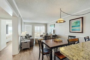 A spacious living area with a dining table, chairs, and a sofa. There is a large window providing natural light and a view of the outdoors. - Pelicans Watch 106 On the Beach (Myrtle Beach)