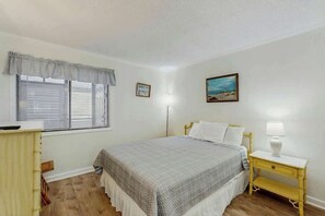 A bedroom with a bed, bedside table, lamp, window with blinds, and a picture on the wall. - Pelicans Watch 106 On the Beach (Myrtle Beach)