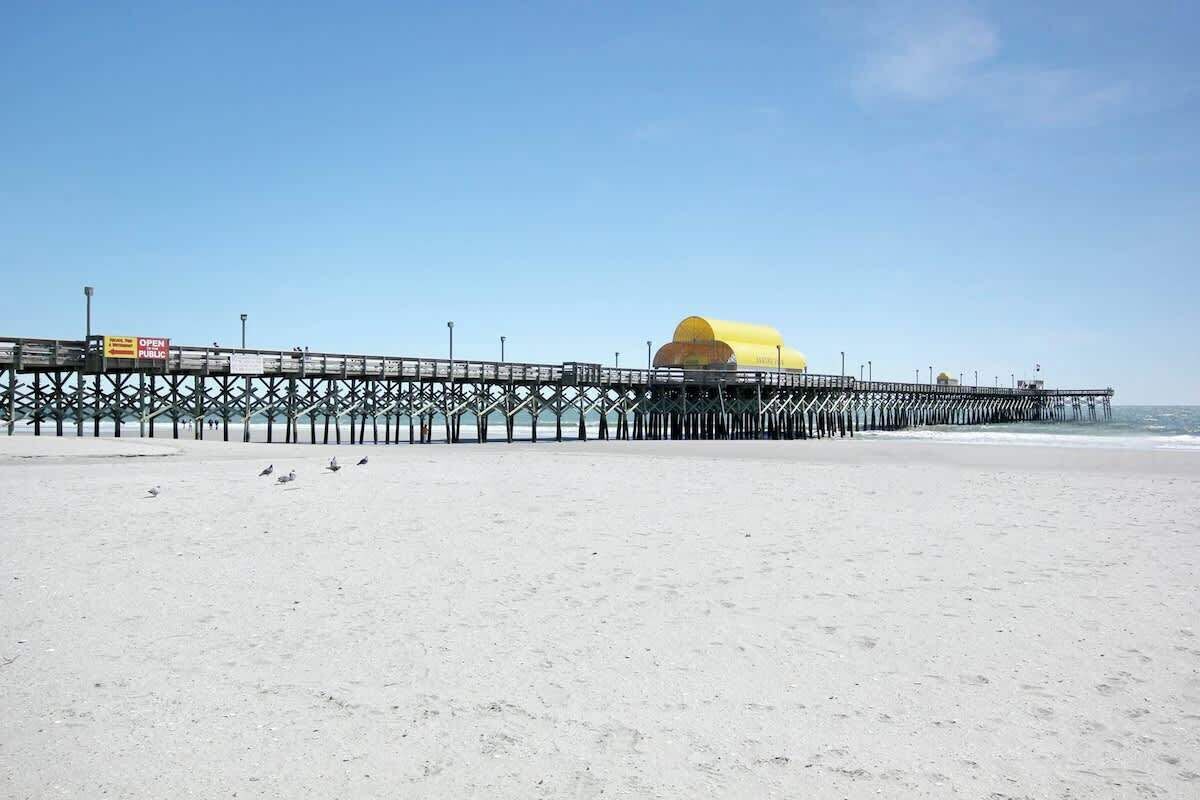 A beach with a pier extending into the water, featuring a yellow structure and a sign that reads 'OPEN PUBLIC.'