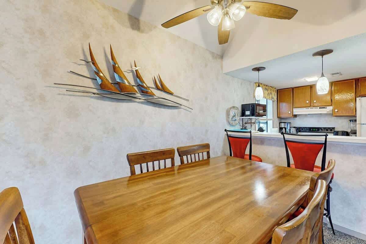 A dining area with a wooden table and chairs, a ceiling fan, and a kitchen in the background.