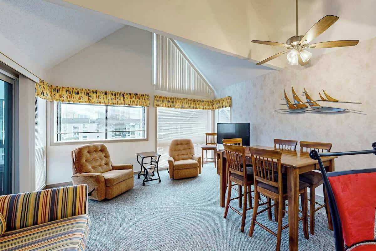 Une pièce comprenant une table à manger, des chaises, un canapé, un ventilateur de plafond et de grandes fenêtres équipées de stores.