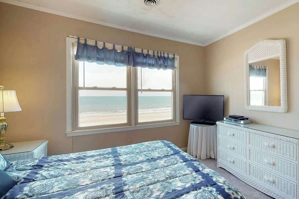 A bedroom with a bed, a television, a dresser, a window with a view of the beach, and a lamp.
