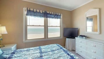 A bedroom with a bed, a television, a dresser, a window with a view of the beach, and a lamp.