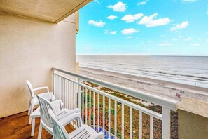 Ein Balkon mit weißen Plastikstühlen, einem Geländer und Blick auf Strand und Meer.
