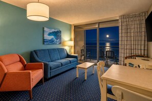 A hotel room with a sofa, armchair, table, and chairs. There is a painting on the wall and a view of the ocean through the window. - The BW | Seven Oh Eight (North Myrtle Beach)