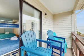 Een balkon met twee blauwe Adirondack-stoelen en een klein tafeltje, een schuifdeur die leidt naar een slaapkamer met een stapelbed, en uitzicht op de oceaan.