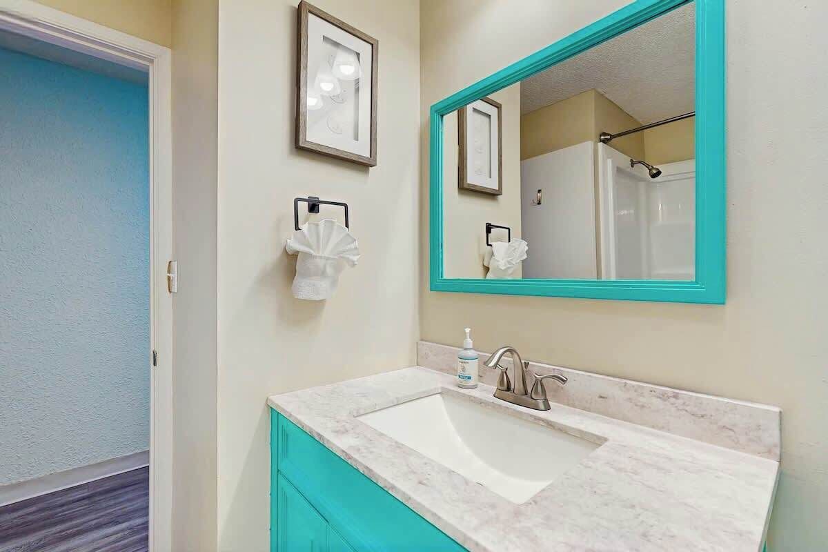 Un baño con un lavamanos blanco, un espejo grande con marco turquesa, y una zona de ducha visible en el reflejo.