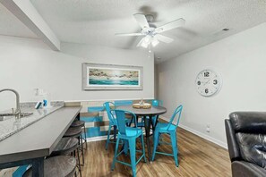 A modern kitchen with a bar area, a dining table with chairs, a ceiling fan, and a clock on the wall. - Sea Turtle Ocean View Condo (Myrtle Beach)