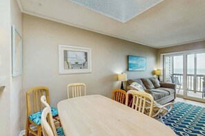 A room with a dining table, chairs, a sofa, a lamp, and a view through a large window. - Atlantic Escape Oceanfront Condo (North Myrtle Beach)