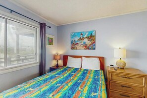 A bedroom with a bed, a nightstand, a lamp, a painting, and a window with blinds. - Atlantic Escape Oceanfront Condo (North Myrtle Beach)