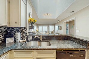 A kitchen with a granite countertop, stainless steel sink, and a dishwasher. There is a window providing natural light and a view of the outdoors. - Atlantic Escape Oceanfront Condo (North Myrtle Beach)