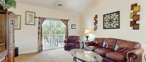 A living room with a leather sofa, a glass coffee table, a ceiling fan, and a view through a glass door to an outdoor patio.