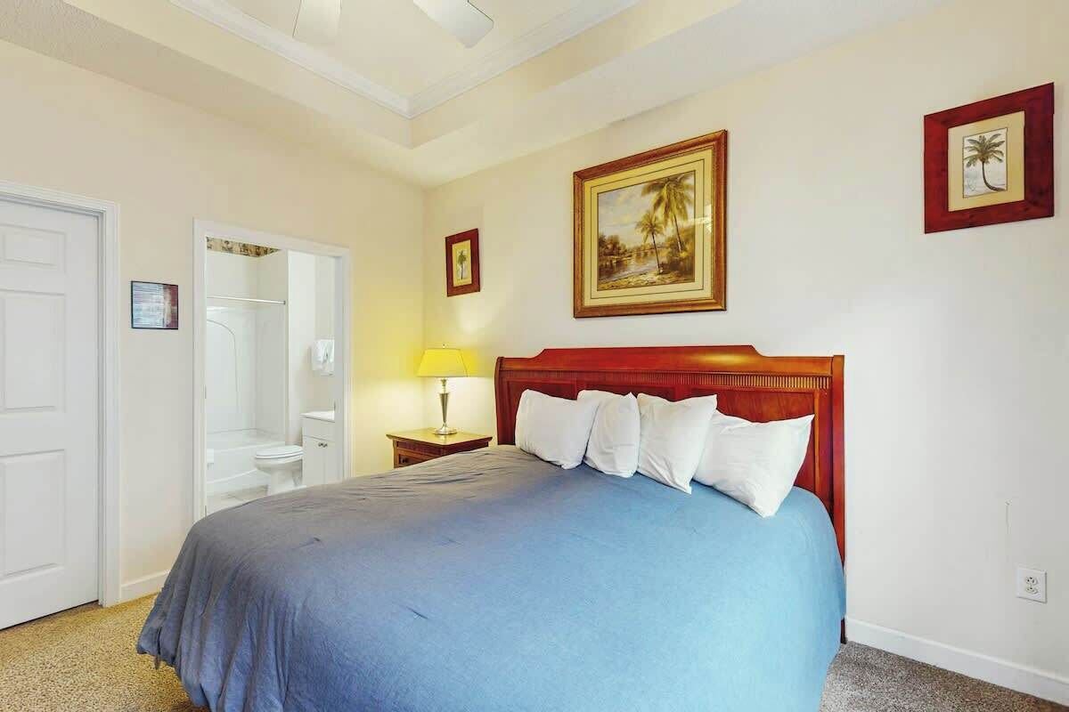 A bedroom with a bed, a nightstand, a lamp, a door, and a wall with framed pictures.