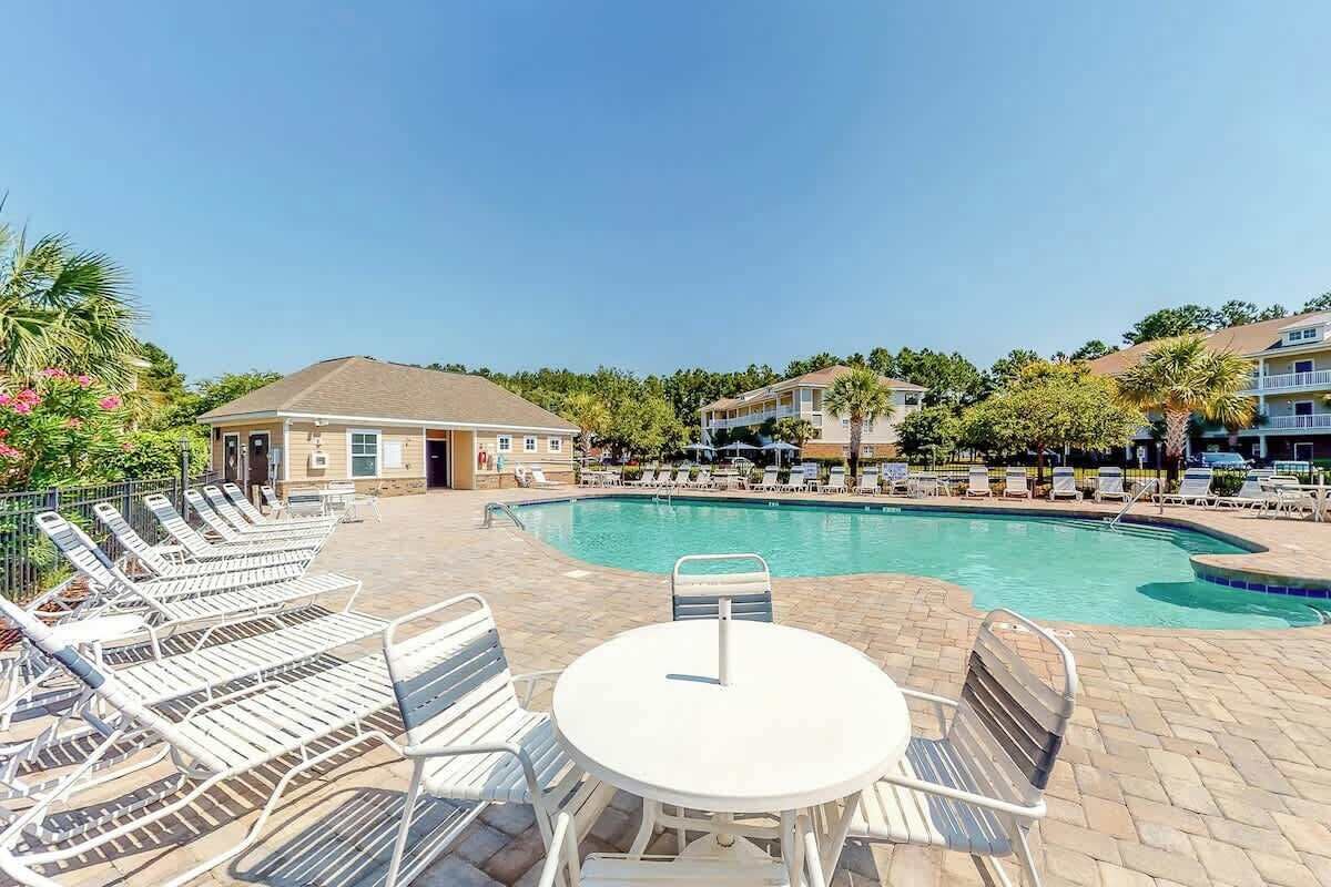 A pool area with lounge chairs and a table, surrounded by buildings and trees.