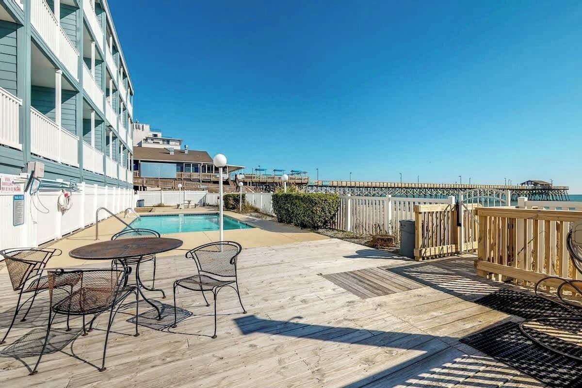 A pool area with a wooden deck, metal chairs, and a view of the pier.