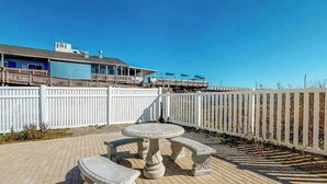 A patio with a round table and benches, a white fence, and a building with a balcony in the background. - Beachfront Blessing Oceanfront Condo (Garden City Beach)