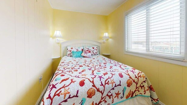 A bedroom with a bed, two bedside lamps, a window with blinds, and a yellow wall. - Beachfront Blessing Oceanfront Condo (Garden City Beach)