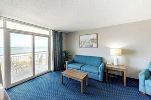 A hotel room with a large window, a sofa, a coffee table, a lamp, and a framed picture on the wall. - Bay Watch Resort 0314 (North Myrtle Beach)