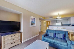 Ein Wohnzimmer mit Flachbildfernseher, einer Couch, einem Couchtisch und einer sichtbaren Küchenzeile im Hintergrund.