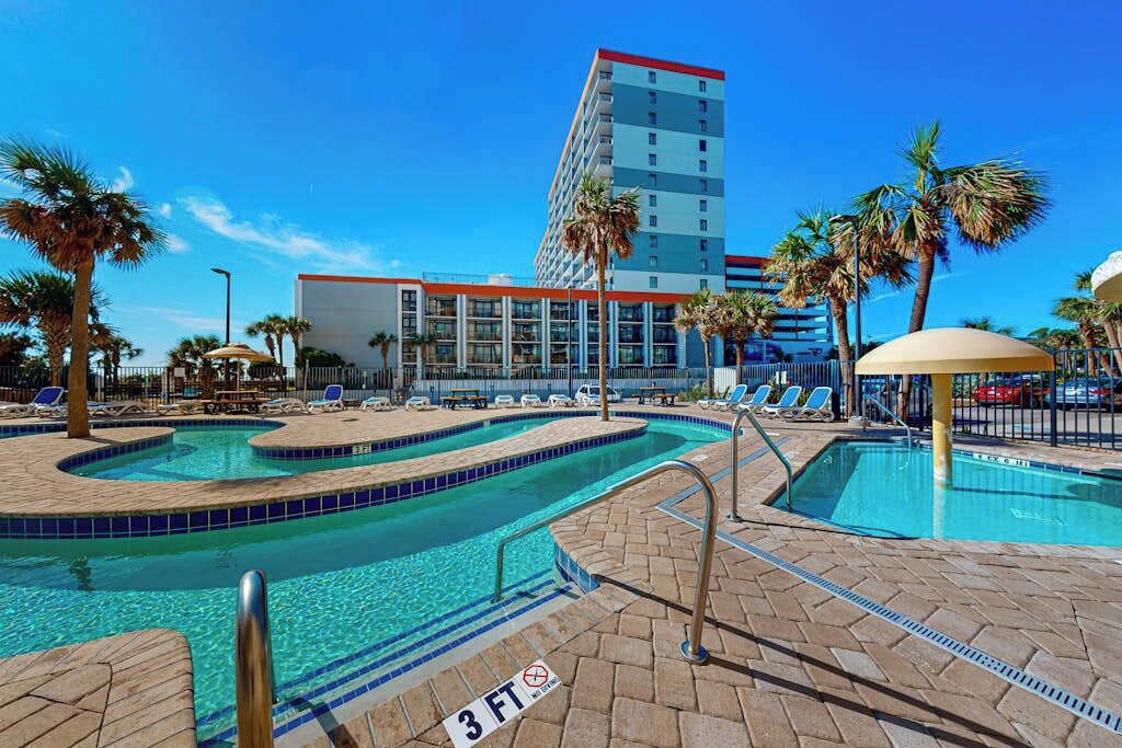 A hotel pool area with multiple swimming pools, palm trees, and a building in the background.
