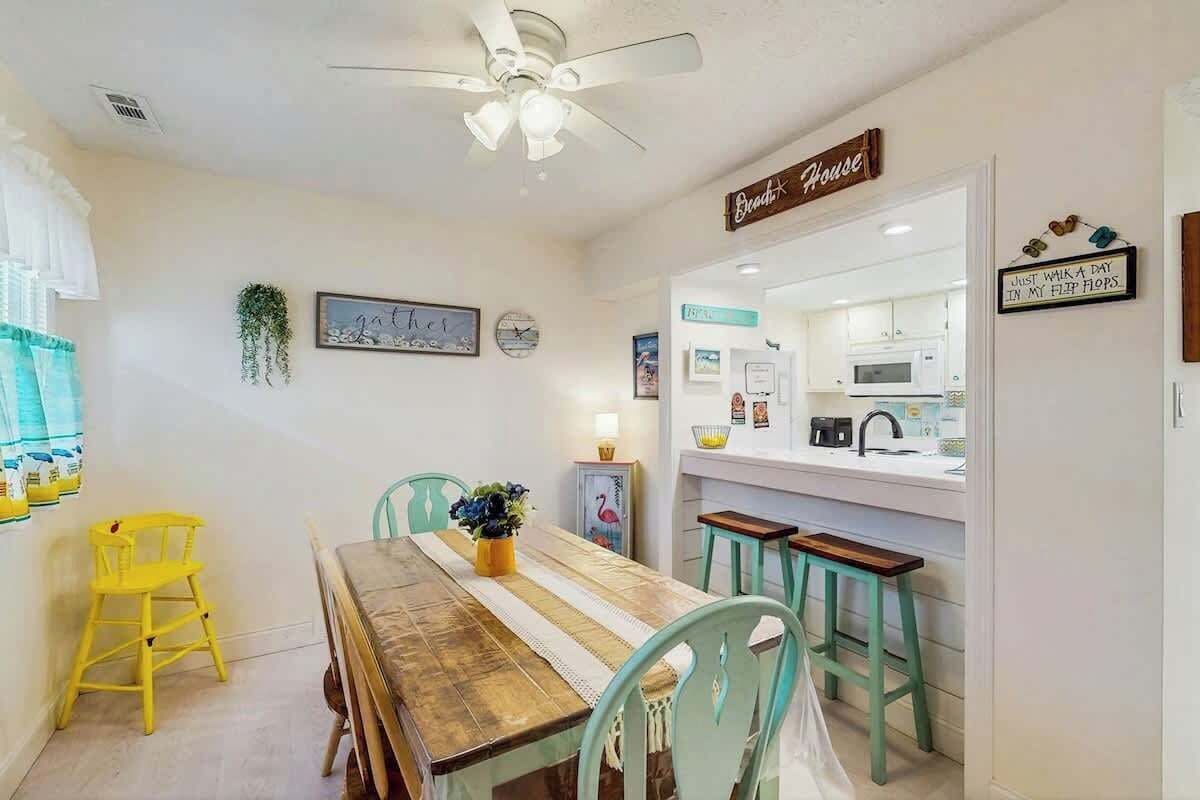 A dining area with a wooden table, chairs, a ceiling fan, and a kitchen visible through an open doorway.