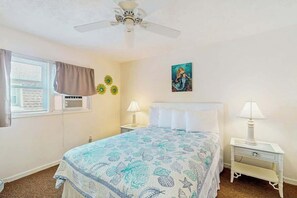 Une chambre à coucher comprenant un lit, deux tables de chevet avec des lampes, un ventilateur de plafond, une fenêtre avec des rideaux et un élément de décoration murale.