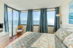 Quarto de hotel com uma cama grande, uma cadeira, uma escrivaninha e vista para o mar através de amplas janelas.