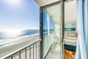 Uma varanda com porta de vidro que dá para um quarto com cama e pintura do oceano.