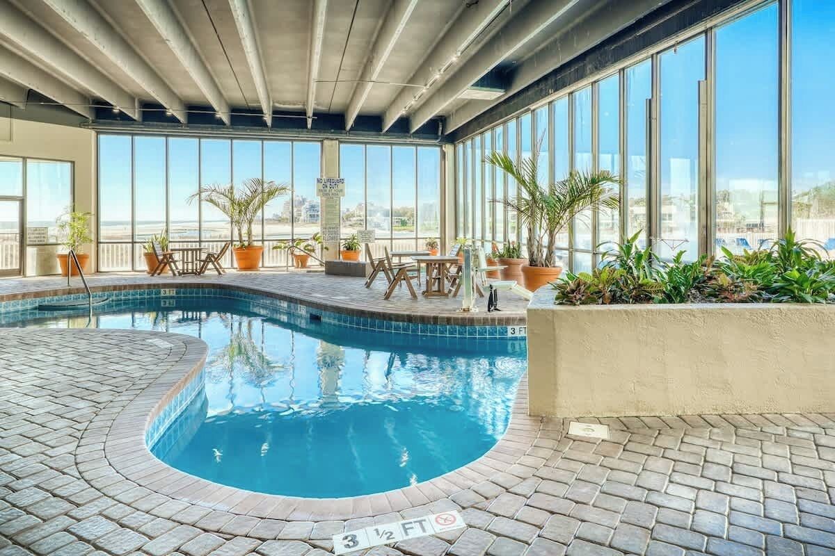 Una piscina interior con suelo de baldosas, rodeada de plantas en macetas y mobiliario de exterior.