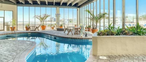 Una piscina interior con suelo de baldosas, rodeada de plantas en macetas y mobiliario de exterior.