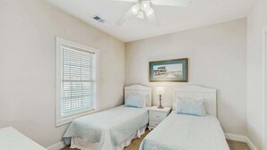 A bedroom with two single beds, a ceiling fan, a window with blinds, a bedside table with a lamp, and a framed picture on the wall. - Magnolia North Unit 304 (Myrtle Beach)
