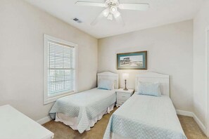 A bedroom with two single beds, a ceiling fan, a window with blinds, a bedside table with a lamp, and a framed picture on the wall. - Magnolia North Unit 304 (Myrtle Beach)