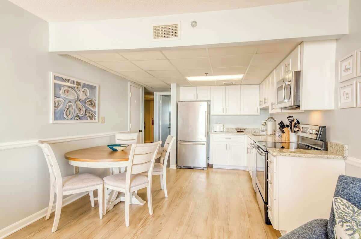 Una cocina compacta con muebles blancos, un refrigerador de acero inoxidable y un comedor con una mesa redonda y sillas.