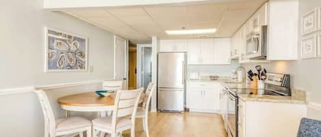 Una cocina compacta con muebles blancos, un refrigerador de acero inoxidable y un comedor con una mesa redonda y sillas.