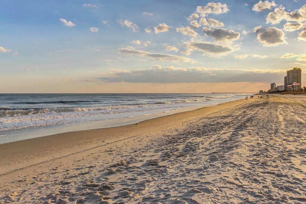 A beach with a clear sky, a pier in the distance, and buildings along the shore.