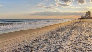 Ein Strand mit blauem Himmel, einer Mole in der Ferne und Gebäuden entlang des Ufers.