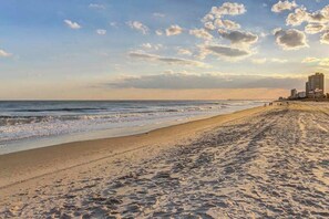 Ein Strand mit blauem Himmel, einer Mole in der Ferne und Gebäuden entlang des Ufers.